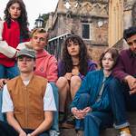 "Six young people sit and pose on historic stone steps outside an old church building, dressed in colorful casual outfits, creating a moody and thoughtful atmosphere in a picturesque outdoor setting."